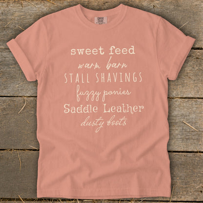 Peach t-shirt with white text: sweet feed, warm barn, stall shavings, fuzzy ponies, saddle leather, dusty boots.