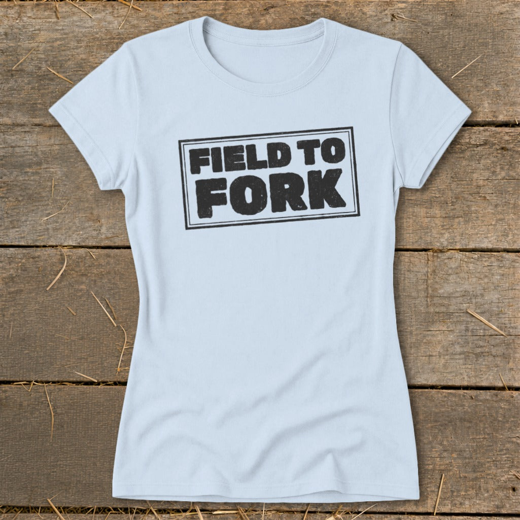 Light blue women's t-shirt featuring the boxed black text FIELD TO FORK, laid flat on a rustic wooden background.
