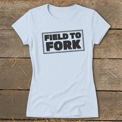 Light blue women's t-shirt featuring the boxed black text FIELD TO FORK, laid flat on a rustic wooden background.