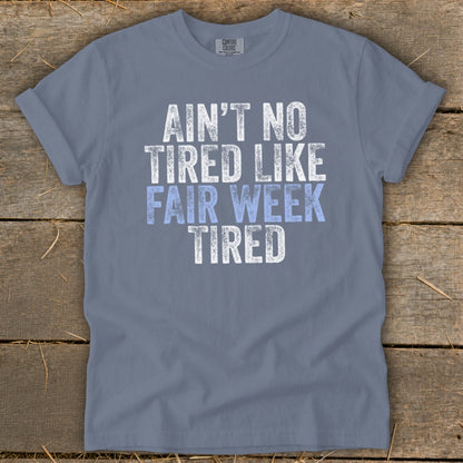 Blue t-shirt with distressed white and light blue text reading "Ain't no tired like fair week tired".