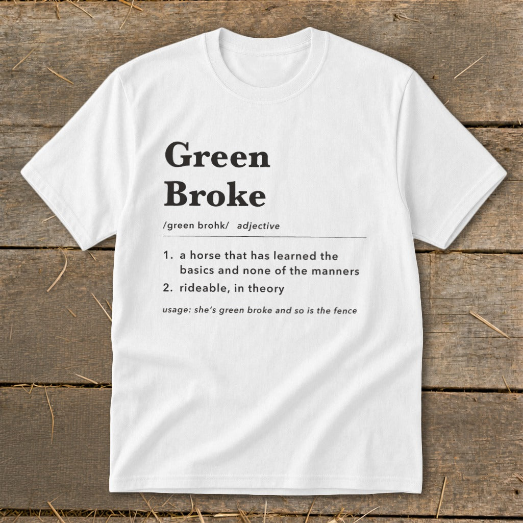 A white t-shirt featuring dictionary-style text defining the term green broke laid flat on a wooden background.