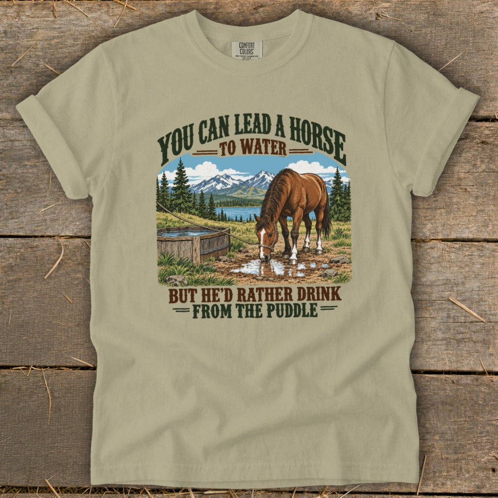 An olive green t-shirt with a graphic of a horse drinking from a puddle with mountains and text.