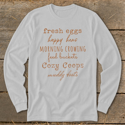 White long-sleeve shirt with "fresh eggs happy hens morning crowing feed buckets cozy coops muddy boots" text.