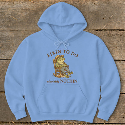 Light blue hoodie with cartoon frog relaxing in rocking chair, text "FIXIN TO DO absolutely NOTHIN".