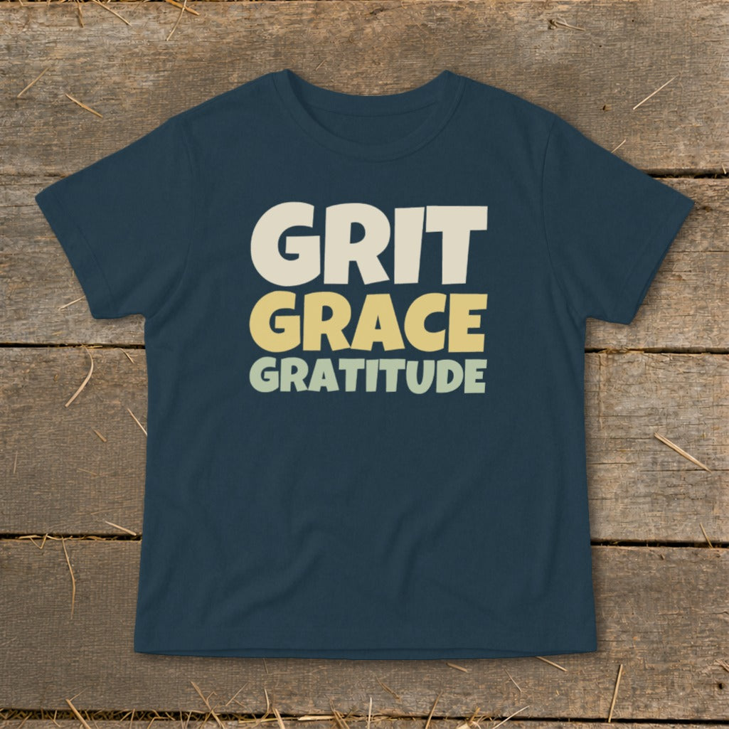 Dark blue t-shirt featuring the stacked white and yellow text GRIT GRACE GRATITUDE, laid flat on a wooden surface.