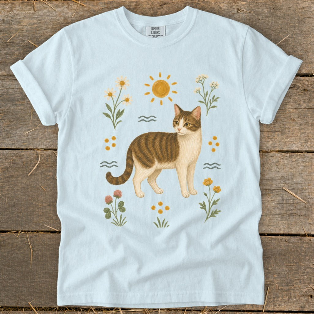 Light blue t-shirt with a brown and white cat surrounded by flowers and sun.