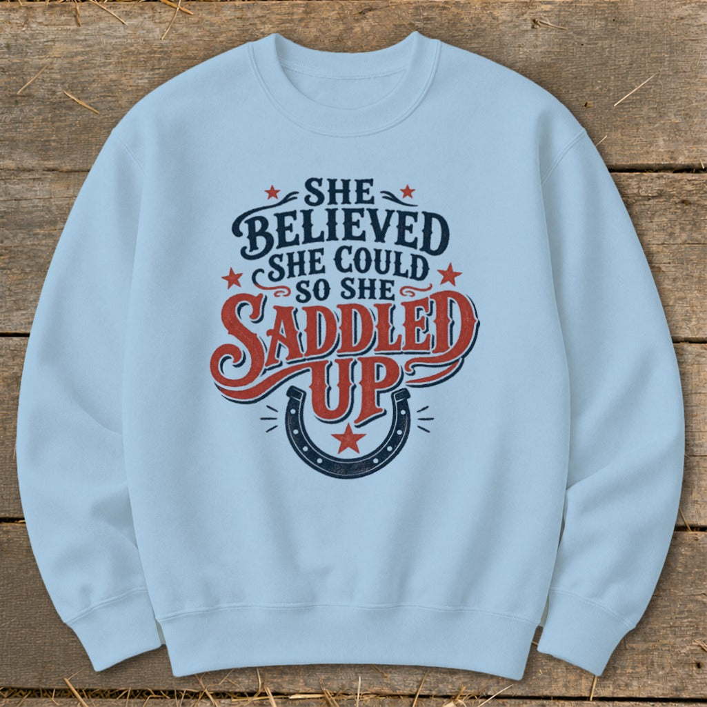 Light blue sweatshirt with "She believed she could so she saddled up" text and horseshoe graphic.
