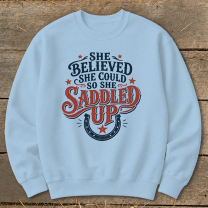 Light blue sweatshirt with "She believed she could so she saddled up" text and horseshoe graphic.