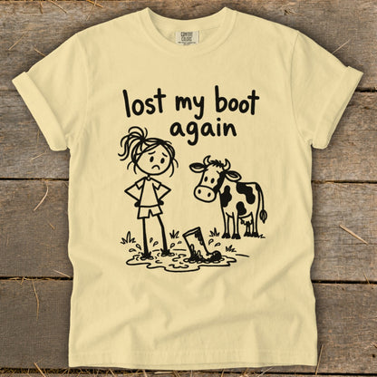 A light yellow t-shirt with a cartoon graphic of a sad girl and a cow standing in mud, and text that reads lost my boot again.