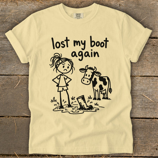 A light yellow t-shirt with a cartoon graphic of a sad girl and a cow standing in mud, and text that reads lost my boot again.