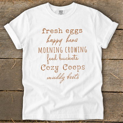 White t-shirt with brown text: fresh eggs, happy hens, morning crowing, feed buckets, Cozy Coops, muddy boots.