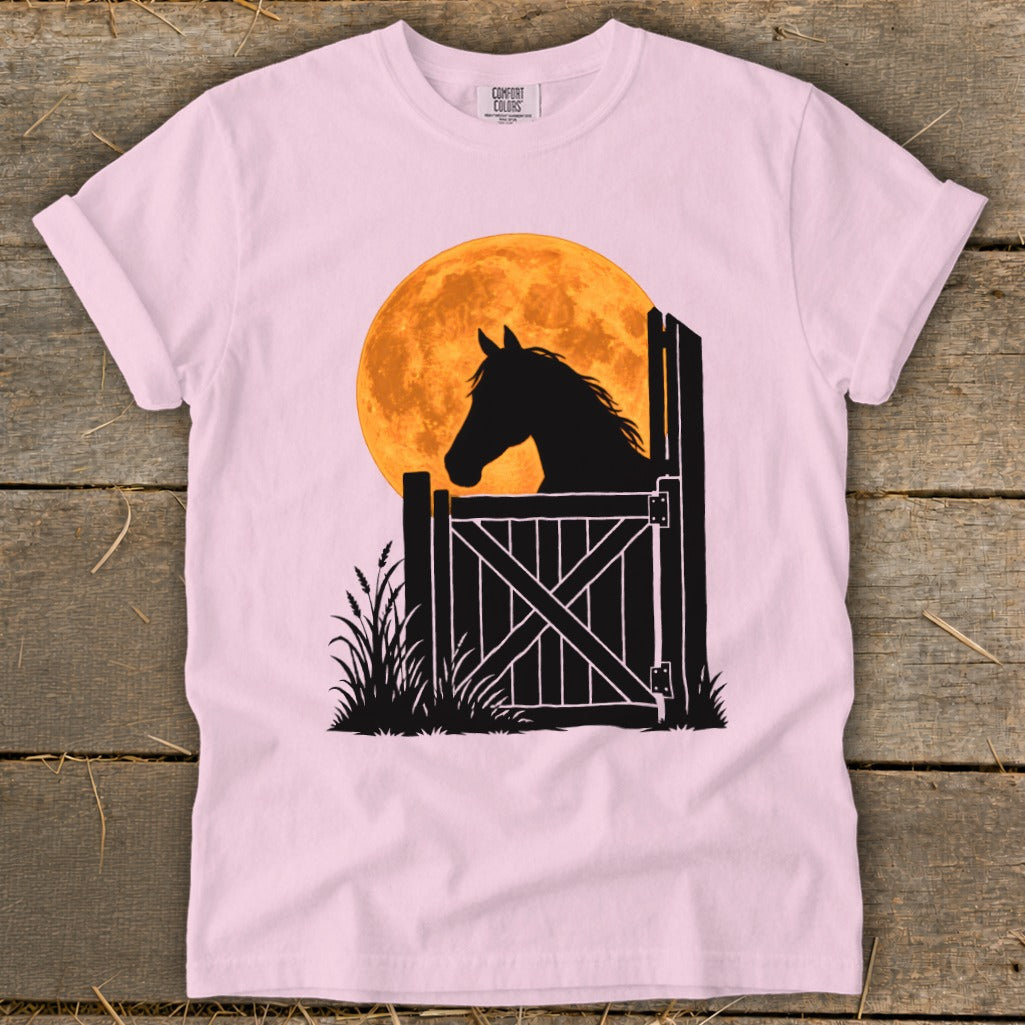 Light pink t-shirt featuring a graphic of a horse's silhouette looking over a gate before a large orange moon.
