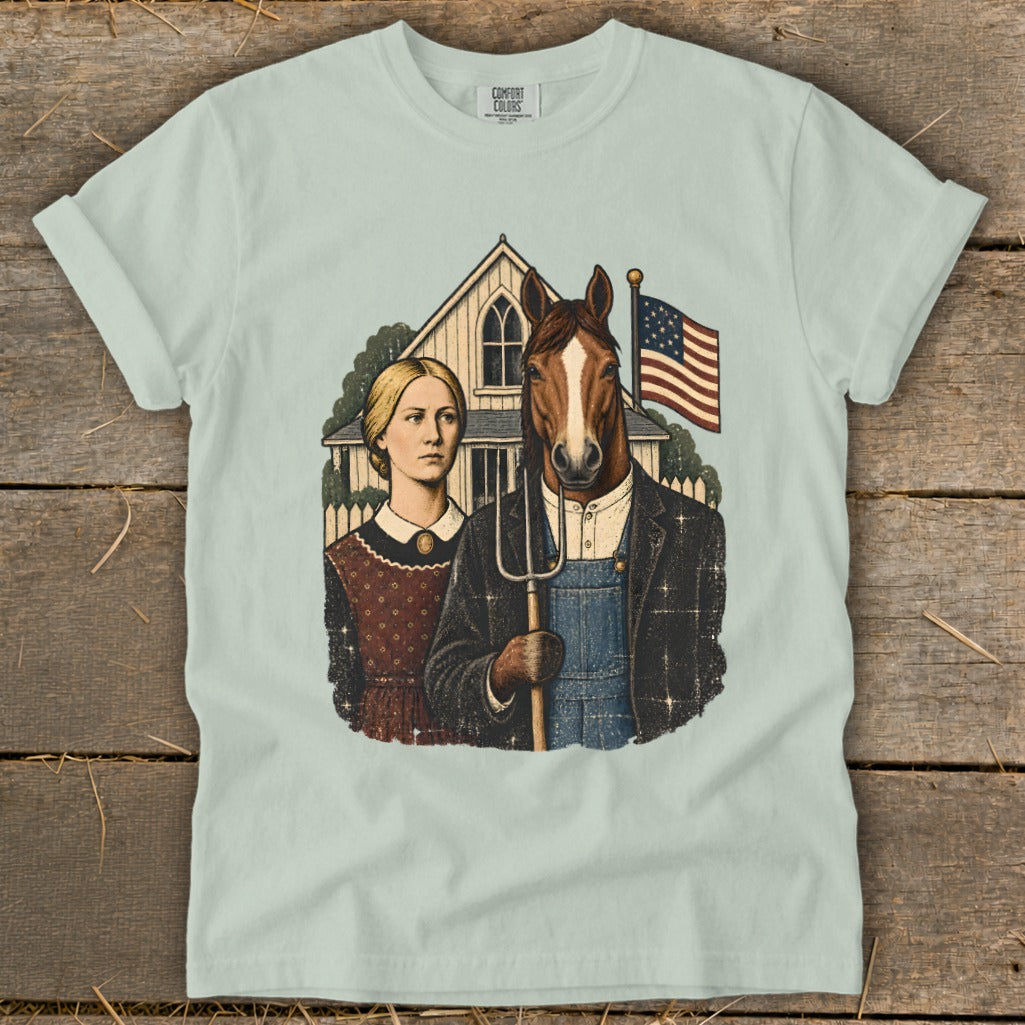 Light green t-shirt with a vintage graphic of a woman and a horse holding a pitchfork in front of an American Gothic style house and American flag.