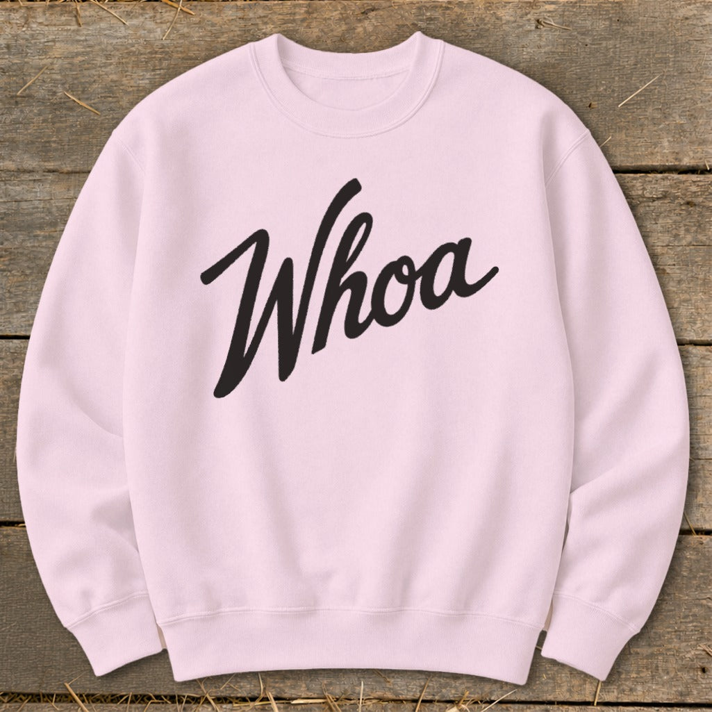 Pink crewneck sweatshirt with black "Whoa" text, laid flat on wood and straw.