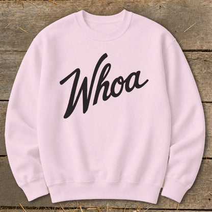 Pink crewneck sweatshirt with black "Whoa" text, laid flat on wood and straw.