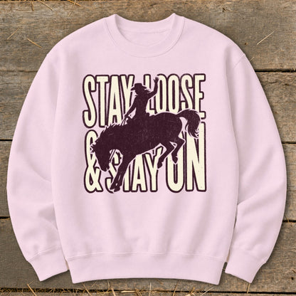 Pink sweatshirt with distressed "STAY LOOSE & STAY ON" text and cowboy silhouette on horse.