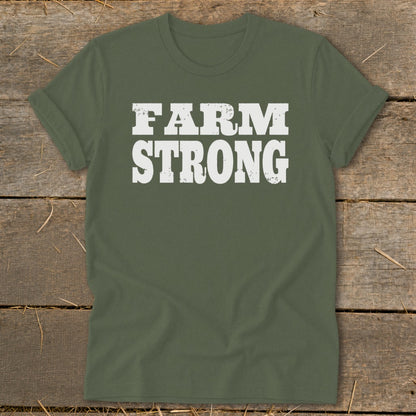 An olive green crewneck t-shirt with distressed white text reading FARM STRONG, displayed flat on a wooden surface.