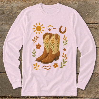 Cowboy boots surrounded by sun, horseshoe, flowers, and leaves on a pink long-sleeve shirt.