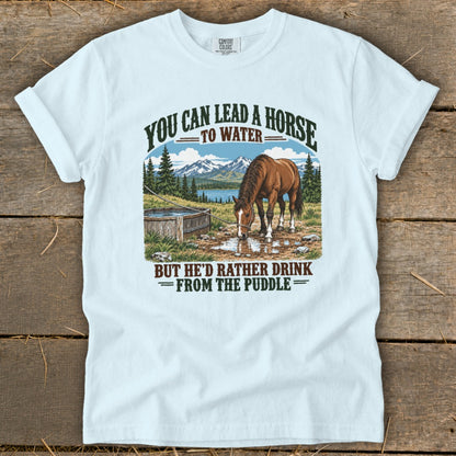 Light blue t-shirt featuring a graphic of a horse drinking from a puddle with mountains and the text You Can Lead a Horse to Water But He'd Rather Drink From the Puddle.