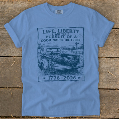 Blue t-shirt with black graphic of man napping in truck bed, reading "Life, Liberty and the Pursuit of a Good Nap in the Truck".