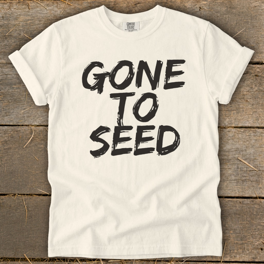 Cream t-shirt with black text "GONE TO SEED" on a wooden background.