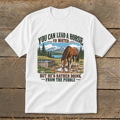 A white t-shirt featuring graphic text and an illustration of a brown horse drinking from a puddle near a wooden trough, with mountains and trees in the background.