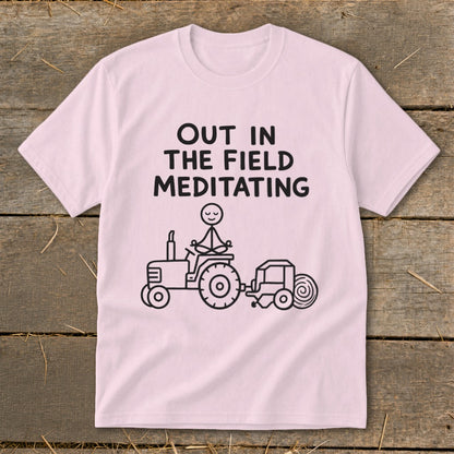 Pink t-shirt with black text "OUT IN THE FIELD MEDITATING" and a stick figure on a tractor.