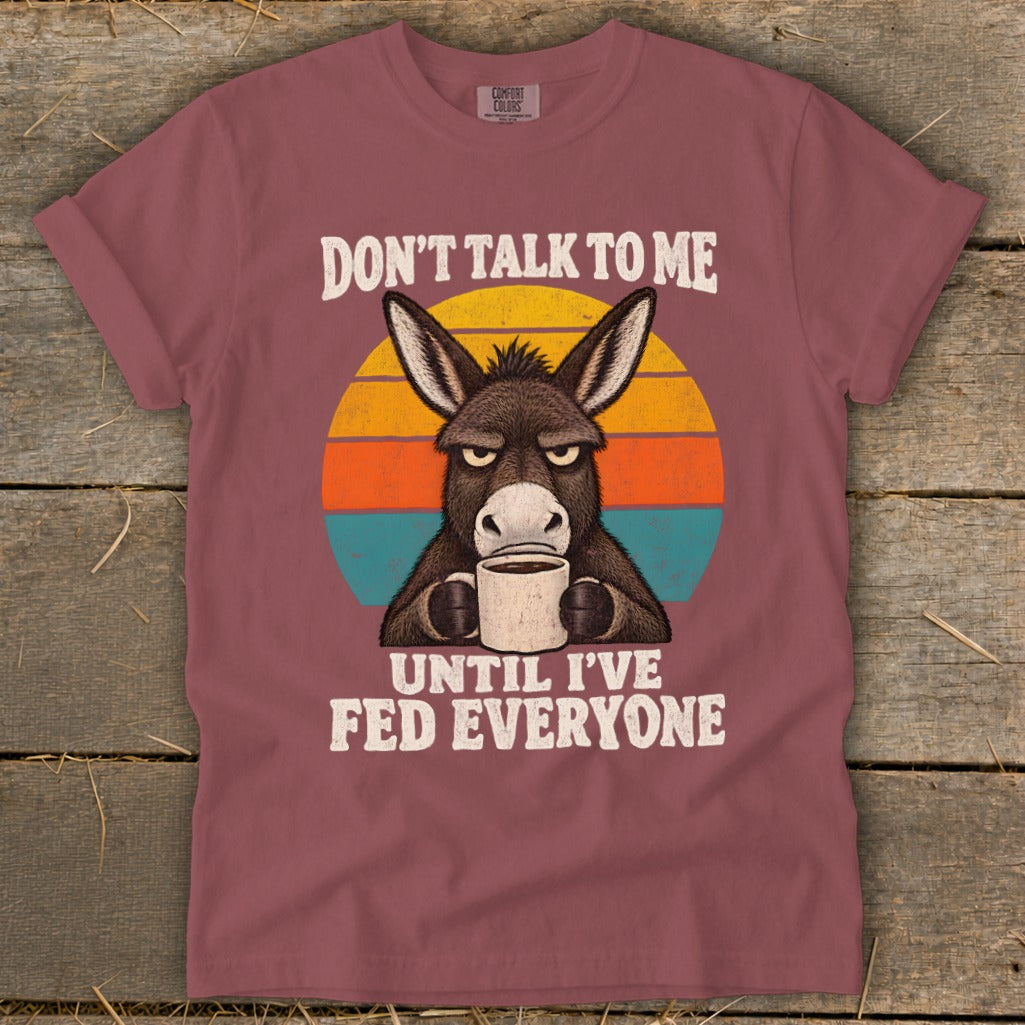Muted red t-shirt with a graphic of an annoyed donkey drinking coffee against a retro sunset.