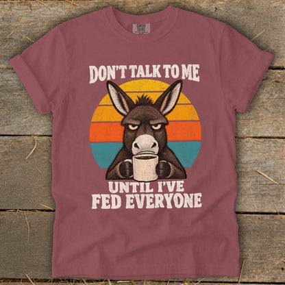 Muted red t-shirt with a graphic of an annoyed donkey drinking coffee against a retro sunset.