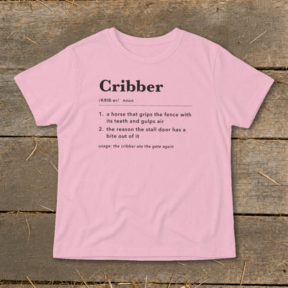 A light pink t-shirt printed with a dictionary definition of the word Cribber, laid flat on a wooden surface.