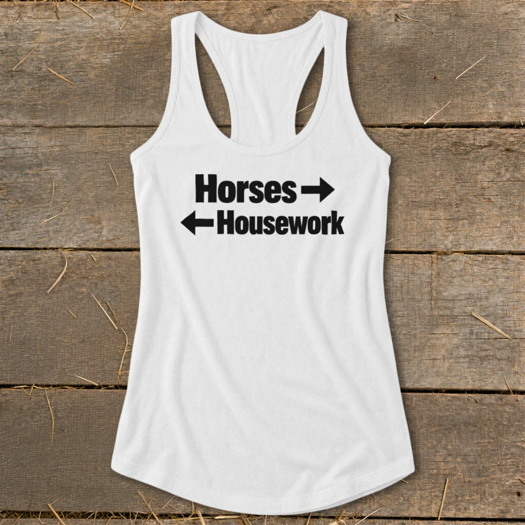 A white racerback tank top with black text reading Horses with a right arrow and Housework with a left arrow, laid flat on a rustic wooden background.