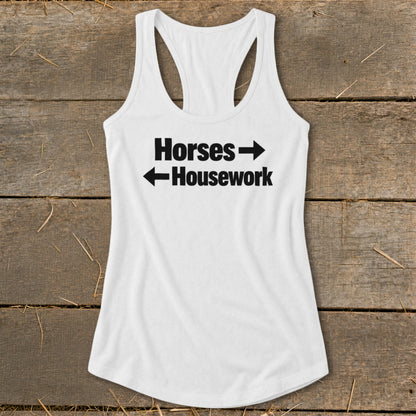 A white racerback tank top with black text reading Horses with a right arrow and Housework with a left arrow, laid flat on a rustic wooden background.