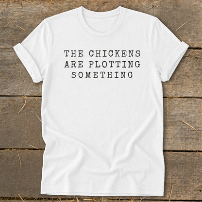 White t-shirt with black text that reads "THE CHICKENS ARE PLOTTING SOMETHING."