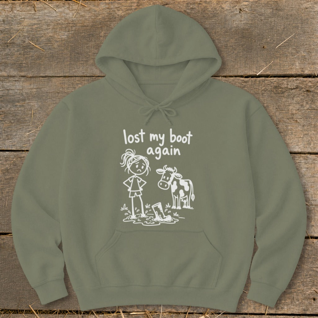 An olive green hoodie featuring a white cartoon graphic of a cow and a girl looking down at a boot stuck in mud, with the text lost my boot again above them.