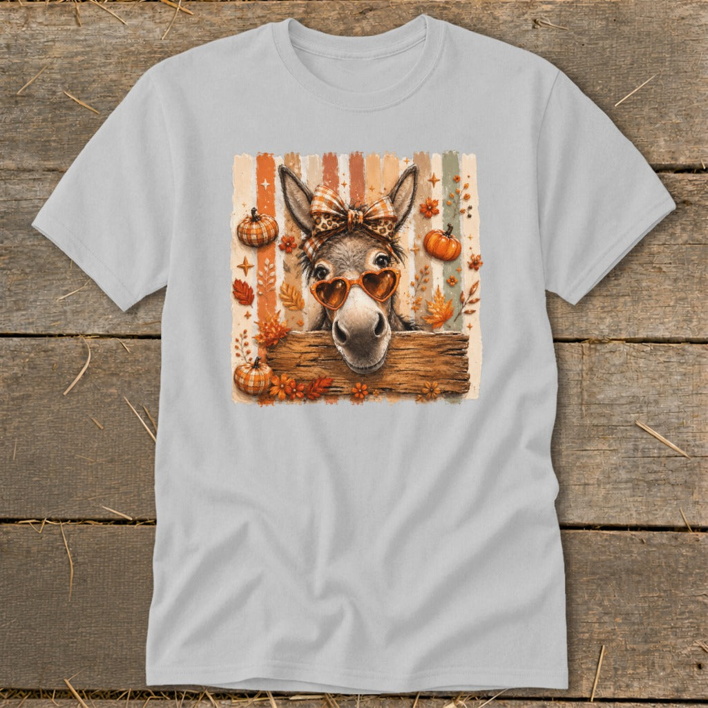 Donkey wearing heart sunglasses and bow peeking over wooden fence, with pumpkins and leaves.