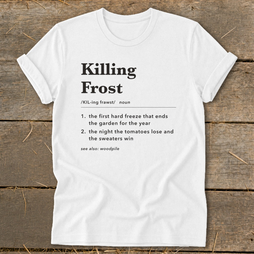 A white t-shirt with black text defining the term Killing Frost, laid flat on a rustic wooden background.