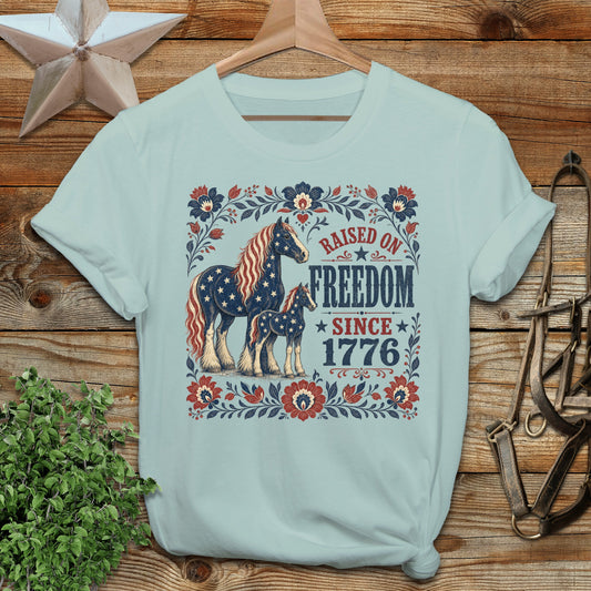 Light blue t-shirt with horses, stars, and "Raised on Freedom Since 1776" text.