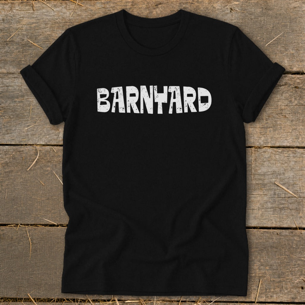 A black crew neck t-shirt with rolled sleeves lies flat on a rustic wooden surface, featuring the word BARNYARD printed in distressed white capital letters across the chest.
