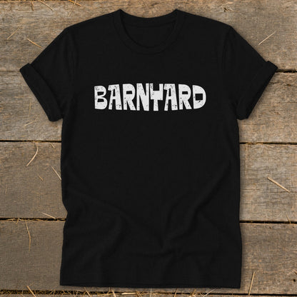 A black crew neck t-shirt with rolled sleeves lies flat on a rustic wooden surface, featuring the word BARNYARD printed in distressed white capital letters across the chest.