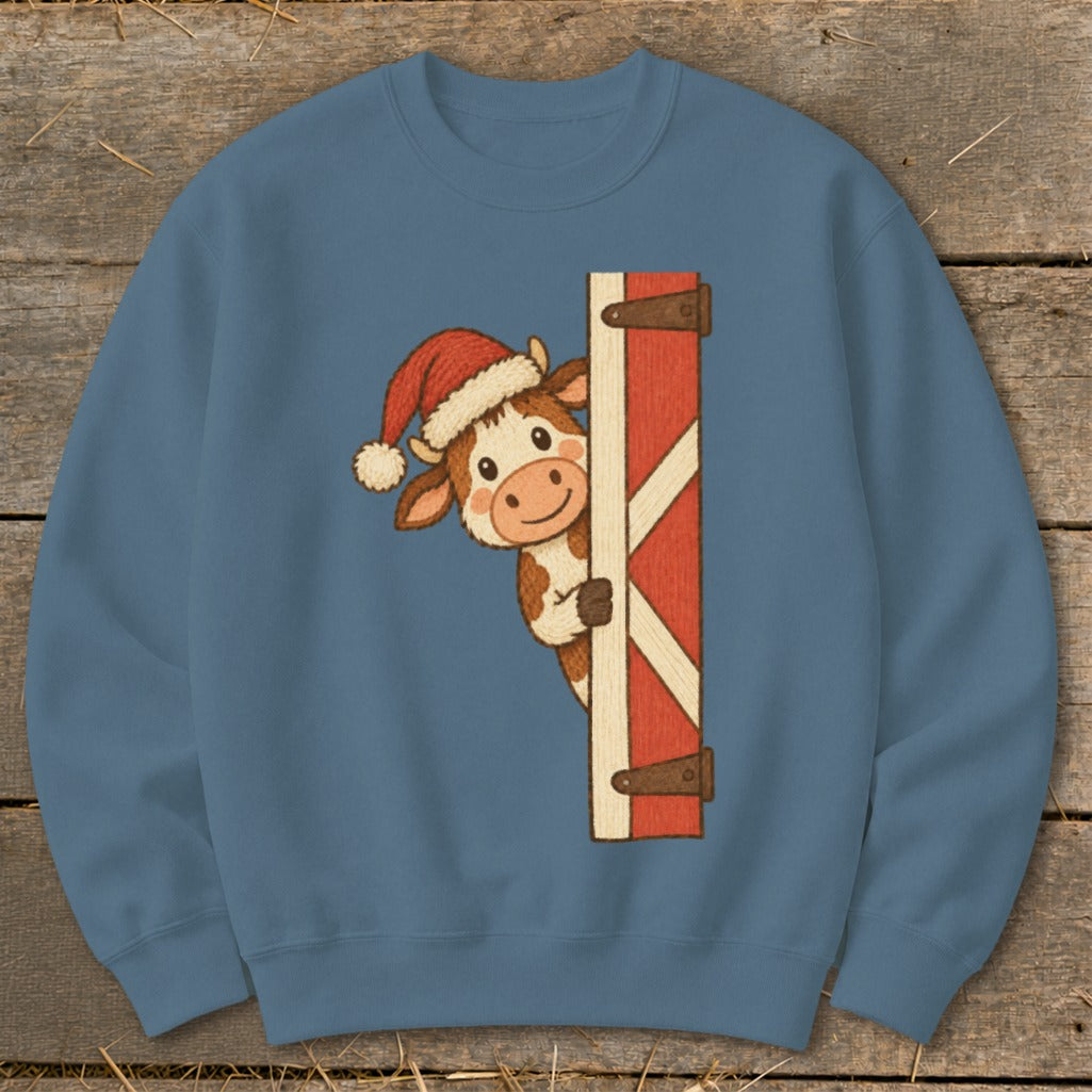 Cow wearing Santa hat peeking around a red barn door on a blue sweatshirt.