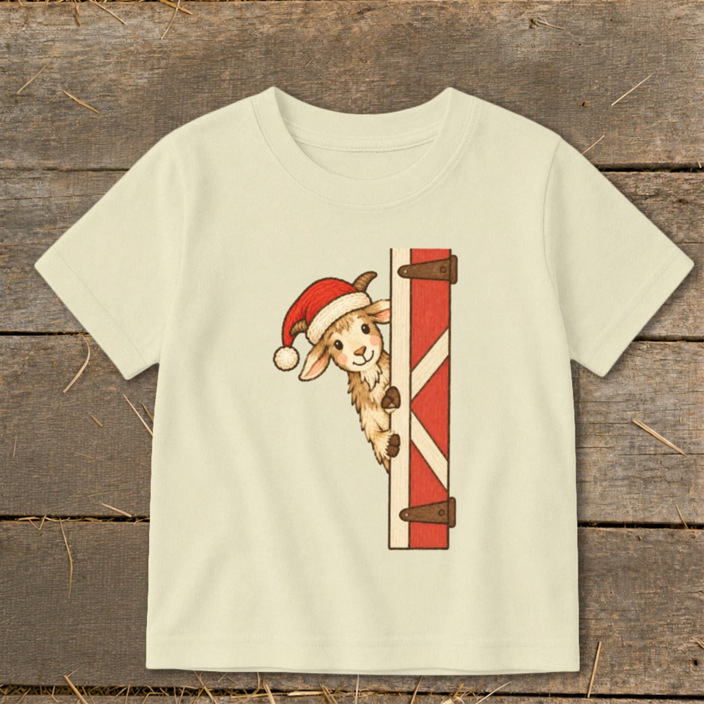 Cream t-shirt with a cartoon goat wearing a Santa hat peeking from behind a red barn door.