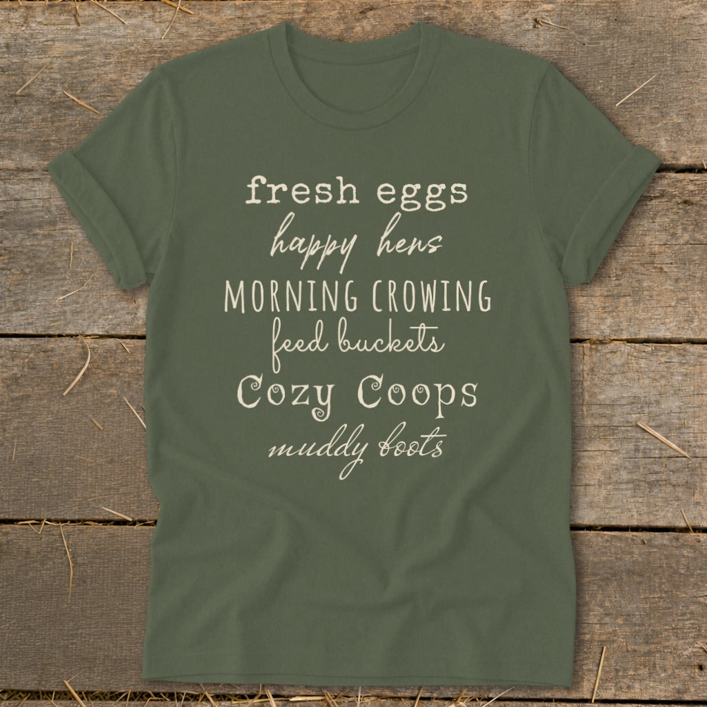 Olive green t-shirt with text: fresh eggs, happy hens, morning crowing, feed buckets, cozy coops, muddy boots.