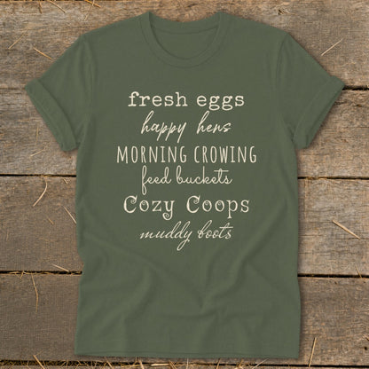 Olive green t-shirt with text: fresh eggs, happy hens, morning crowing, feed buckets, cozy coops, muddy boots.