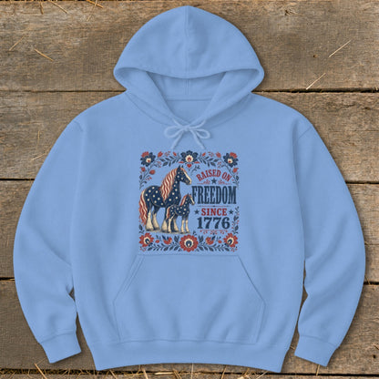 Light blue hoodie with a distressed graphic of two horses and text "RAISED ON FREEDOM SINCE 1776".