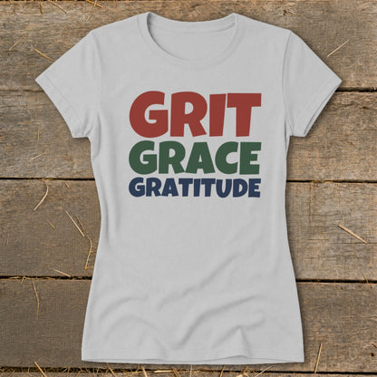 A light gray t-shirt featuring the stacked text Grit, Grace, and Gratitude lies on wood.