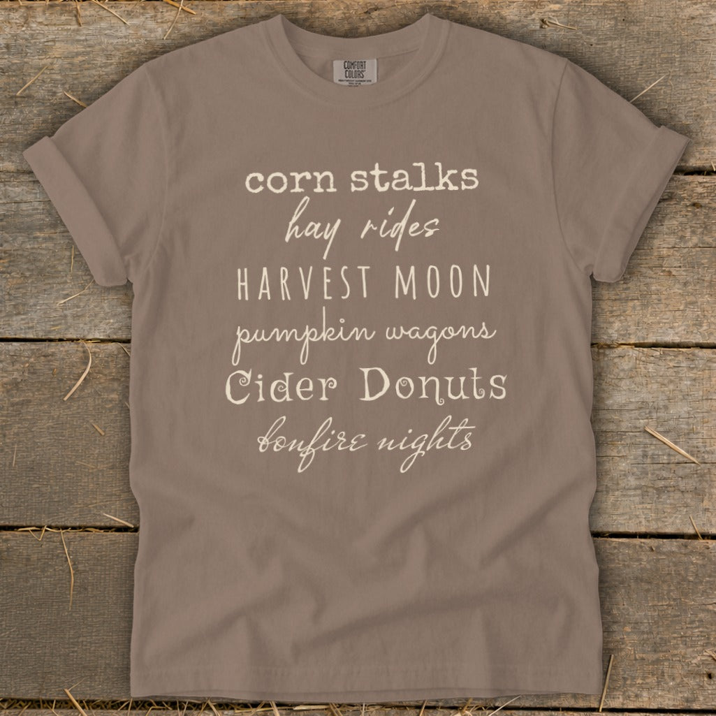 Brown t-shirt with white text listing fall activities: corn stalks, hay rides, harvest moon, pumpkin wagons, cider donuts, bonfire nights.