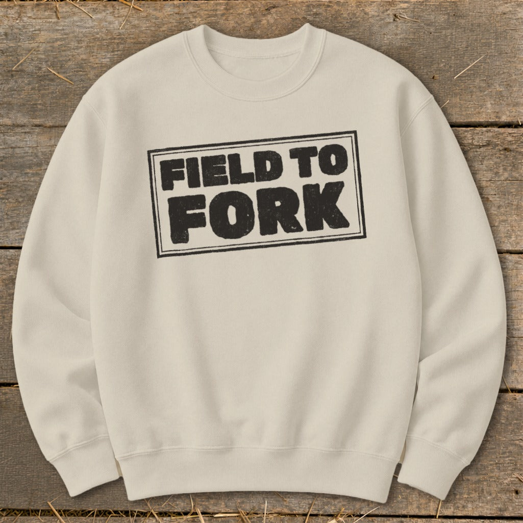 Cream-colored crewneck sweatshirt featuring a rectangular black graphic with the text FIELD TO FORK centered on a rustic wooden background.