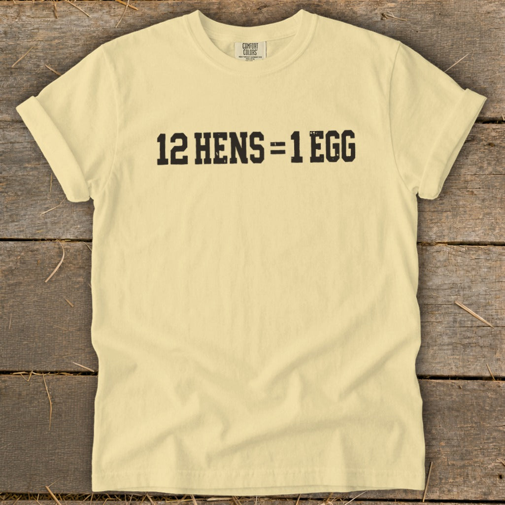 A pale yellow t-shirt laying flat on a rustic wooden surface displays the black text 12 HENS = 1 EGG.