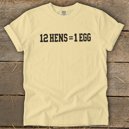 A pale yellow t-shirt laying flat on a rustic wooden surface displays the black text 12 HENS = 1 EGG.