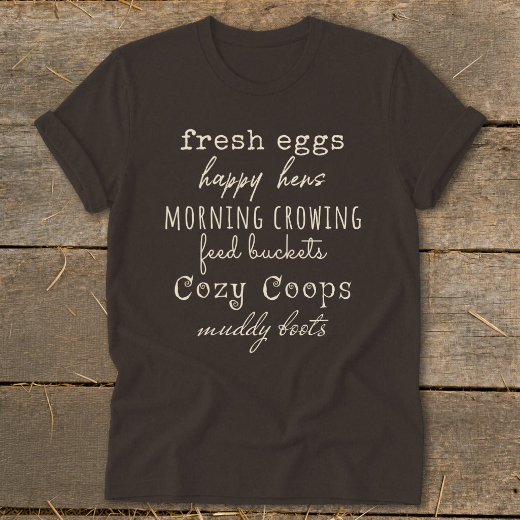 Dark t-shirt with white text listing farm-related phrases: fresh eggs, happy hens, morning crowing, feed buckets, cozy coops, muddy boots.
